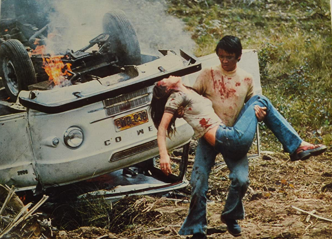 zdjęcie: mężczyzna w zakrwawionej koszulce niesie nieprzytomną kobietę, w tle znajduję się przewrócony, płonący pojazd