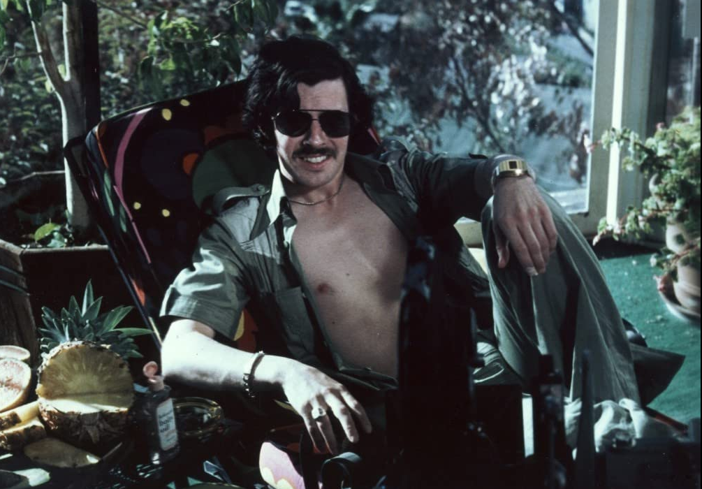zdjęcie: uśmiechnięty siedzący mężczyzna z wąsem, ubrany w rozpiętą koszule i okulary przeciw słoneczne