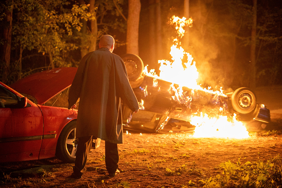 zdjęcie: odwrócony tyłem łysy mężczyzna w płaszczu, w tle przewrócony płonący samochód w śród drzew